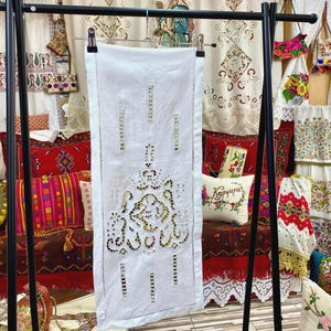 May include: A white linen table runner with intricate cutwork and a decorative floral design. The runner is hanging on a black metal rack, showcasing its detailed craftsmanship. Other embroidered textiles are visible in the background.