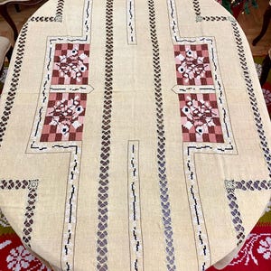 May include: A beige oval tablecloth with intricate embroidered designs in brown, pink, and white. The tablecloth features a repeating pattern of floral motifs and geometric shapes.