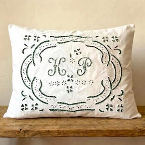 May include: White rectangular pillow with a decorative green embroidered border and the letters "K" and "P" in the center. The pillow is placed on a rustic wooden surface.