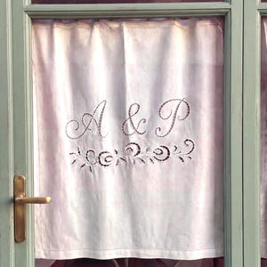 May include: A white fabric curtain panel featuring intricate cutwork embroidery. The centre displays the letters A & P in a cursive font, positioned above a decorative floral vine motif. The curtain is mounted within a light green window frame.