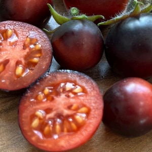 May include: Close-up of several ripe tomatoes, some whole and some halved. The tomatoes are a deep burgundy color with dark purple tops. The cut tomatoes reveal a vibrant red interior with visible seeds. The tomatoes are on a wooden surface.