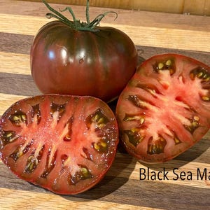 May include: Three Black Sea Man tomatoes, one whole and two halves, on a wooden cutting board. The whole tomato is dark red with green stripes. The cut tomatoes reveal a red interior with visible seeds. The text "Black Sea Man" is visible.