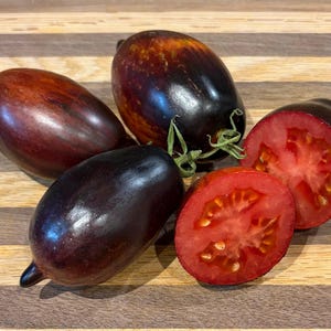 Op de afbeelding: Een assortiment rijpe tomaten, sommige heel en andere gehalveerd, gepresenteerd op een houten oppervlak. De tomaten variëren in kleur, met dieprode en paarse tinten, naast levendig rood. De gesneden tomaten onthullen een sappig interieur.