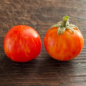 May include: Two round, red and orange striped tomatoes sit on a dark wooden surface. One tomato has a green stem and calyx. The tomatoes have a vibrant, fresh appearance, suggesting they are ripe and ready to eat.