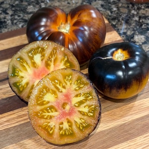 May include: A close-up of several heirloom tomatoes on a wooden cutting board. The tomatoes are a mix of colors, including dark purple, orange, and yellow. Two tomatoes are sliced open, revealing the interior flesh.
