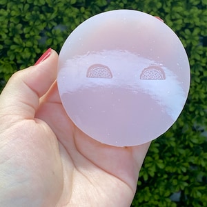 May include: A round, light pink object with two taco designs etched into the surface. The object is held in a hand against a backdrop of green foliage. The tacos are a lighter shade of pink.