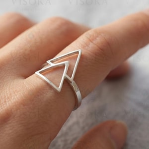 May include: A silver ring featuring a geometric design composed of two open, interconnected triangles. The band is thin and circular, supporting the triangular focal point that sits prominently on the finger. The metal has a smooth, polished finish.