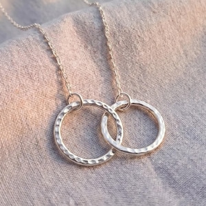 May include: A silver necklace featuring two interlocking circular pendants with a hammered, textured finish. The delicate chain connects to the rings, which are crafted from a reflective metallic material, creating a simple and cohesive design.