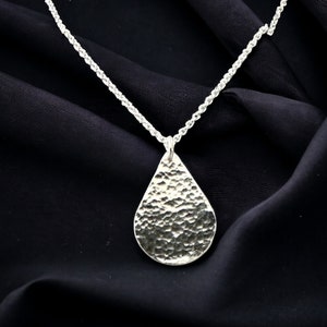 May include: A silver teardrop-shaped pendant necklace with a hammered texture hangs on a silver chain.