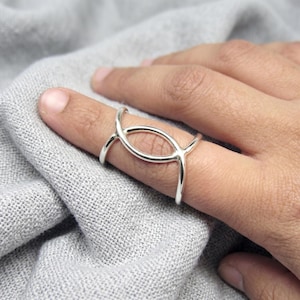 Puede incluir: Un anillo de tono plateado con un acabado liso y pulido, que presenta un diseño abierto de óvalo alargado que rodea el dedo. La banda se divide en dos líneas que convergen en los lados, creando una pieza central simétrica y hueca.