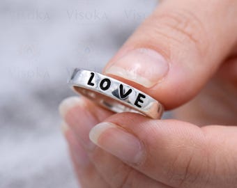 May include: A silver-toned metal ring featuring the word LOVE engraved in black capital letters along the outer band. The ring has a smooth, polished finish and a flat surface profile, showcasing the bold, dark lettering clearly on the band.