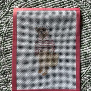 May include: A rectangular needlepoint canvas featuring a printed design of a small bear wearing a red and white striped shirt. The canvas has a white grid pattern and is bordered on all four sides by a solid pink fabric tape binding.