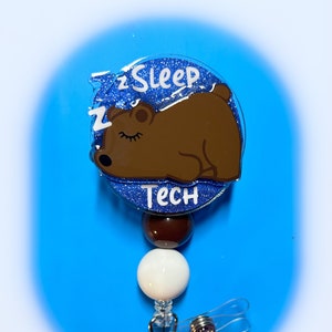 May include: A blue glitter badge holder with a brown bear sleeping on it. The badge holder says "zSleep Tech" and has a white and brown bead attached to a clear retractable badge clip.