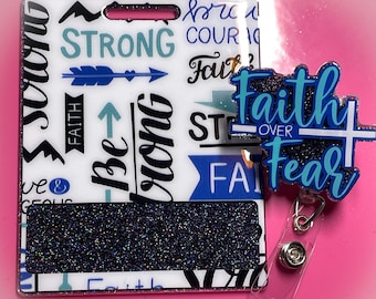 May include: A white and blue glitter badge holder with the words "Strong", "Be Strong", "Faith", "Courage", and "Faith Over Fear" printed on it. The badge holder has a black glitter insert for a name tag. A blue and white glitter charm with the words "Faith Over Fear" is attached to the badge holder.