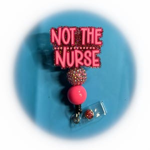 May include: A pink badge reel with the text "NOT THE NURSE" in pink letters. The badge reel has a pink and glittery top, a pink bead, and a clear plastic clip. The background is a light blue colour.