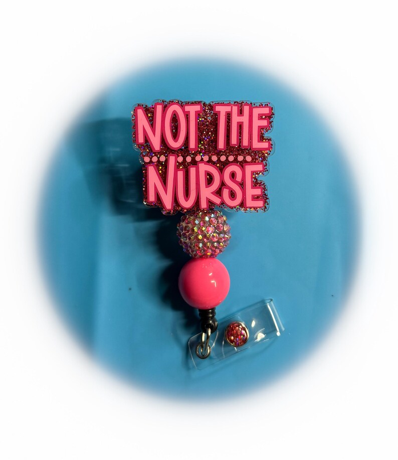 May include: A pink badge reel with the text "NOT THE NURSE" in pink letters. The badge reel has a pink and glittery top, a pink bead, and a clear plastic clip. The background is a light blue colour.