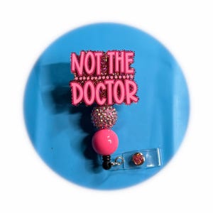 May include: A pink retractable badge reel with the text "NOT THE DOCTOR" in a glittery font. The badge reel has a pink ball and a sparkly accent, with a clear plastic clip.