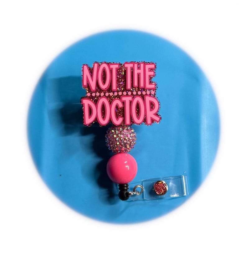 May include: A pink retractable badge reel with the text "NOT THE DOCTOR" in a glittery font. The badge reel has a pink ball and a sparkly accent, with a clear plastic clip.