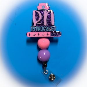 May include: A purple and pink glitter badge reel with a nurse's hat and the text "RN in progress" with a heartbeat graphic. The reel has two beads, one pink and one purple.