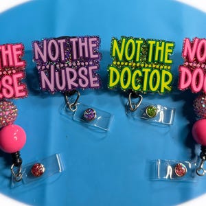 May include: Four badge reels with the phrases "NOT THE NURSE" and "NOT THE DOCTOR" in pink, purple, and green glitter lettering. Each reel has a pink or crystal bead and a clear plastic badge holder.