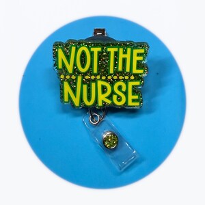 May include: A badge reel displaying the text "NOT THE NURSE" in green lettering with gold glitter detailing. The reel is connected to a clear plastic holder with a green gem. Set against a solid blue circle background.