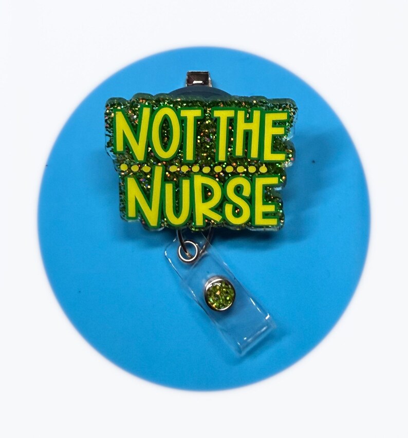 May include: A badge reel displaying the text "NOT THE NURSE" in green lettering with gold glitter detailing. The reel is connected to a clear plastic holder with a green gem. Set against a solid blue circle background.