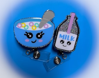Pode incluir: Uma tigela de cereal azul com uma colher e uma caixa de leite branca com um canudo rosa. Ambos têm rostos de desenhos animados fofos e a caixa de leite diz "LEITE". Eles estão presos a um porta-crachá retrátil de plástico transparente.