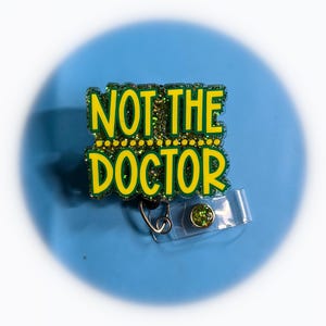 May include: A badge reel with the text "NOT THE DOCTOR" in yellow letters outlined in green glitter. The badge reel has a clear plastic strap and a green gem on the button.