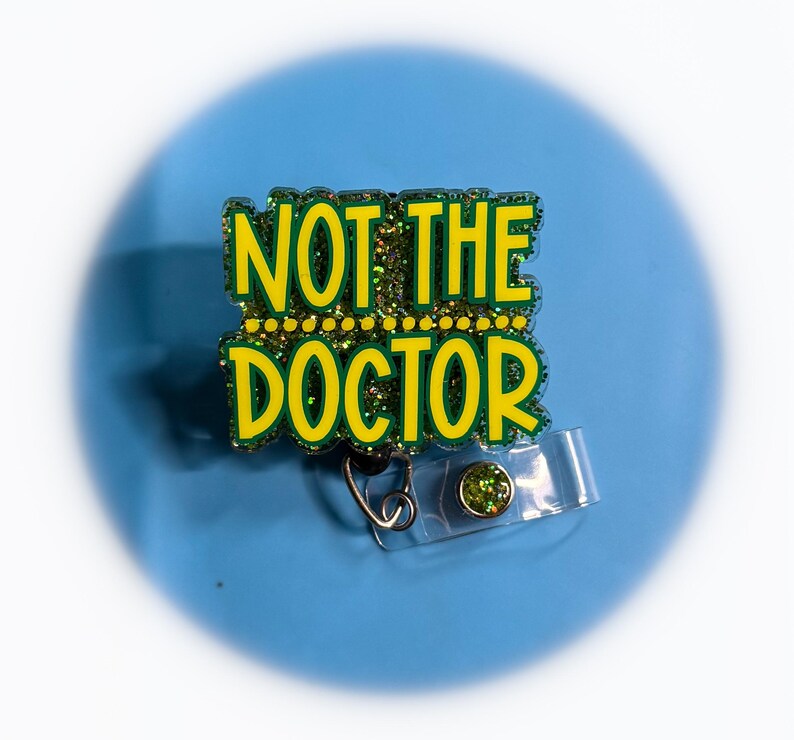 May include: A badge reel with the text "NOT THE DOCTOR" in yellow letters outlined in green glitter. The badge reel has a clear plastic strap and a green gem on the button.