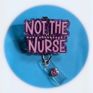 May include: A purple badge reel with the text "NOT THE NURSE" in a bubble letter font. The badge reel has a clear plastic strap and a purple gem on the end. The background is a light blue circle.