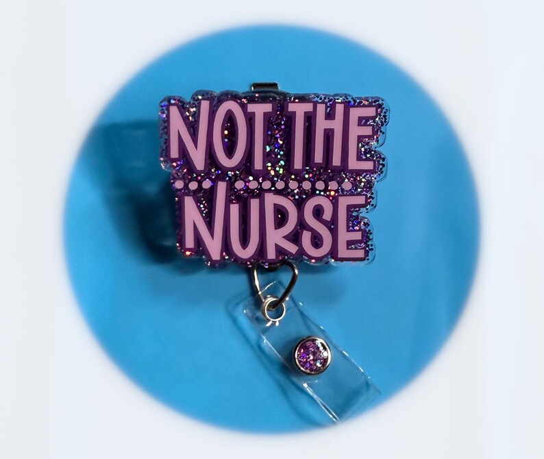 May include: A purple badge reel with the text "NOT THE NURSE" in a bubble letter font. The badge reel has a clear plastic strap and a purple gem on the end. The background is a light blue circle.