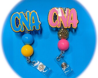 May include: Two colorful retractable badge reels with the word "CNA" in bold letters. One reel has blue and yellow beads, the other has pink and gold beads.
