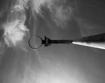 May include: Black and white photograph of a basketball hoop and pole against a cloudy sky. The perspective is from below, looking up at the hoop and the long, cylindrical pole extending into the sky. The sky is filled with wispy clouds.