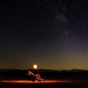 May include: A person reclines in a chair under a starry night sky, illuminated by a warm lamp. The scene is set in a dark, open landscape with a mountain range in the distance. The lamp casts a soft glow.