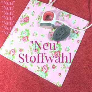 Könnte beinhalten: Eine rosa Stoffmatte mit Blumenmuster und dem Text "Neu" Stoffwahl. Eine kleine weiße und graue Heißpresse mit rotem Griff liegt auf der Matte. Ein schwarzer Heißpressaufsatz liegt ebenfalls auf der Matte.