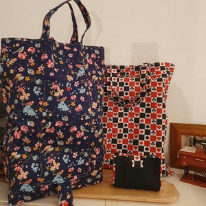 Puede incluir: Dos bolsas de la compra reutilizables. Una bolsa es azul marino con un estampado floral y pájaros. La otra bolsa es de cuadros negros y blancos con corazones y picas rojos.