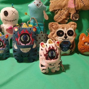 Könnte beinhalten: Eine Sammlung bunter Plüschtiere mit gestickten Details. Die Spielzeuge umfassen ein blaues und rotes Monster, ein weißes und rosa Monster, einen braunen Waschbär und eine orange Katze. Die Spielzeuge sind alle handgefertigt und haben einen skurrilen, cartoonartigen Stil.