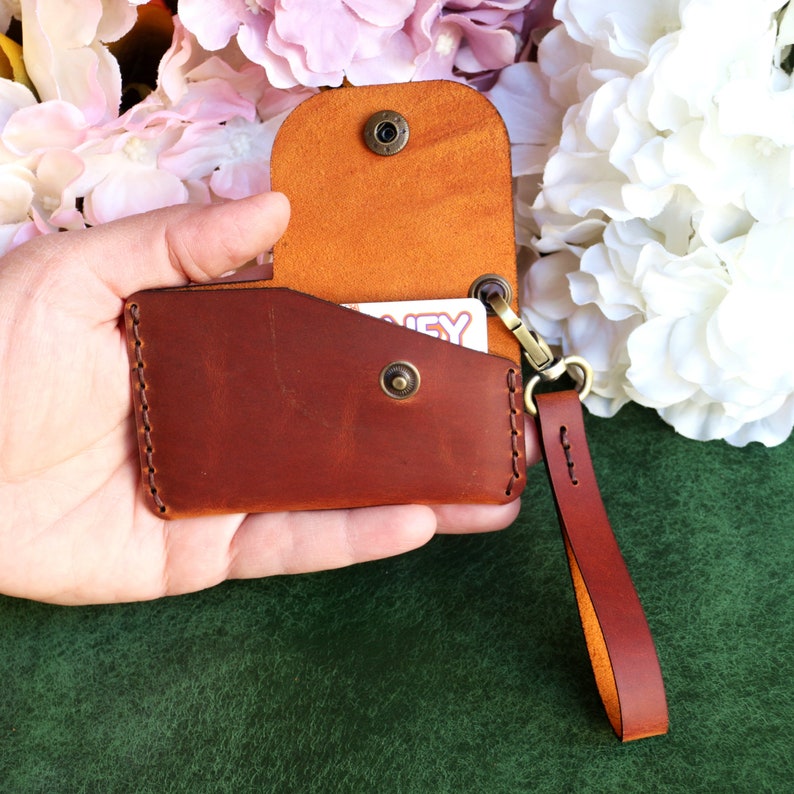 May include: A brown leather wallet with a flap and a strap. The wallet is open and has a card inside. The wallet has a brown leather strap with a metal clasp.