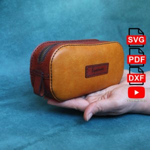 Può includere: Un astuccio in pelle artigianale nei toni del marrone e beige, con chiusura a zip e una piccola etichetta rettangolare. L'astuccio è tenuto in mano, mostrando le sue dimensioni e il suo design. Il testo "Eyeress" è visibile sull'etichetta.