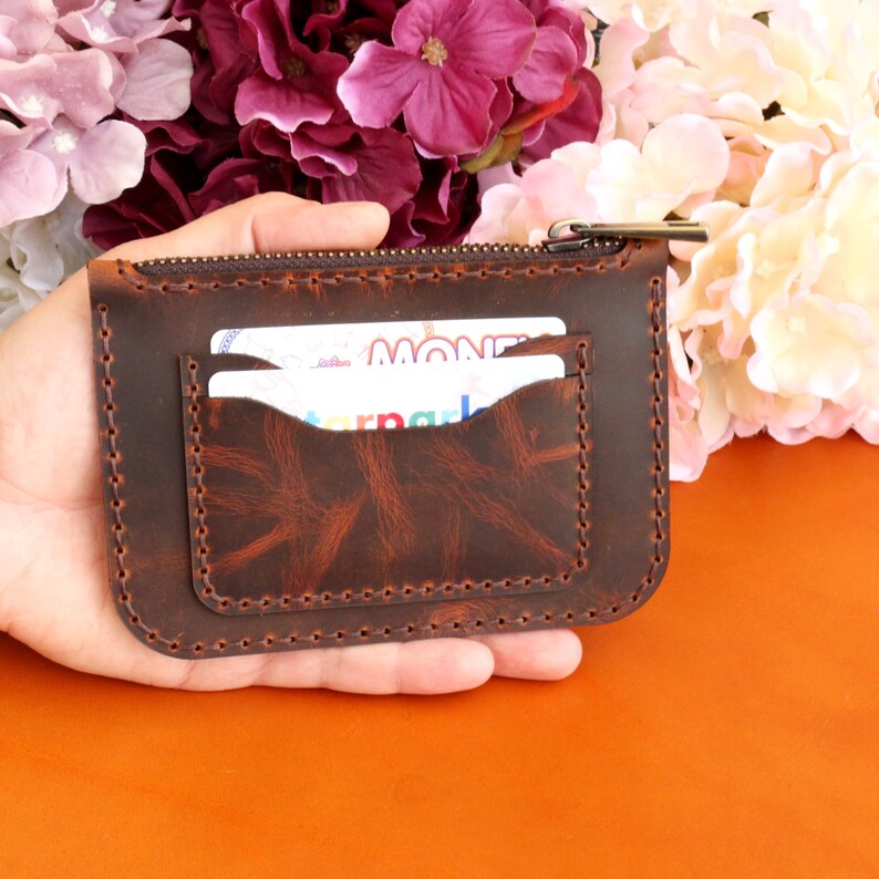 May include: A brown leather wallet with a zipper closure and two card slots. The wallet is held in a hand.