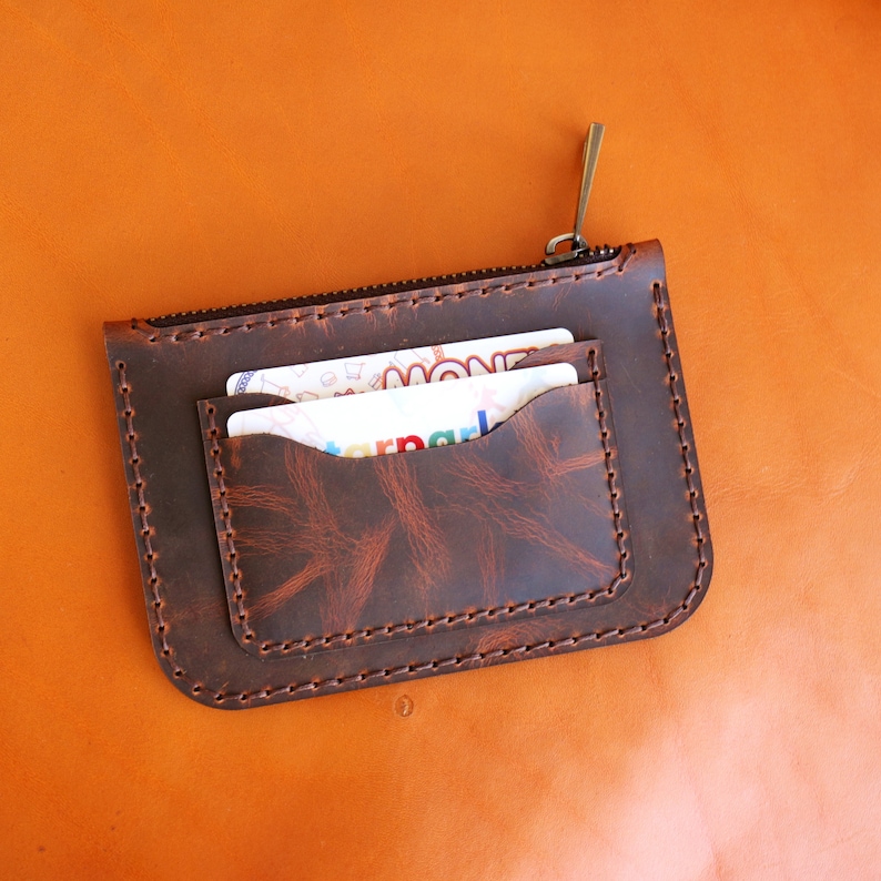 May include: A brown leather zippered wallet with two card slots and a stitched design. The wallet is closed and has a zipper pull.