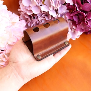 May include: A brown leather holder with three holes for storing items, such as pens or pencils. The holder has a stitched edge and a metal rivet on the side.