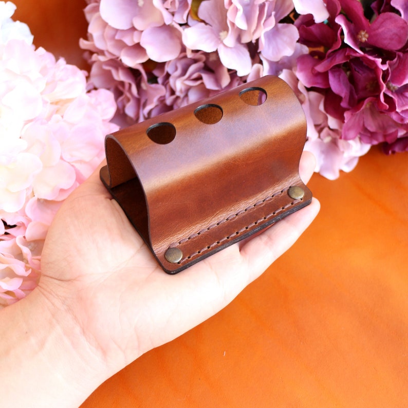 May include: A brown leather holder with three holes for storing items, such as pens or pencils. The holder has a stitched edge and a metal rivet on the side.