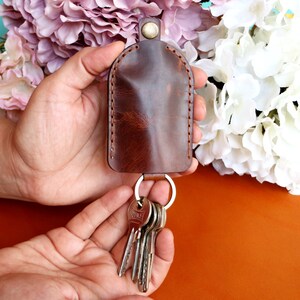 May include: A brown leather key holder with a stitched edge and a silver key ring with keys attached. The key holder is held in a hand.