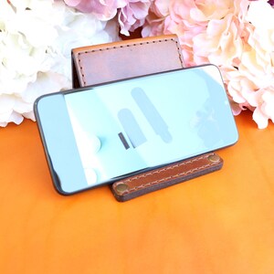 Puede incluir: Un soporte de tel&eacute;fono de cuero marr&oacute;n con un tel&eacute;fono inteligente encima. El soporte tiene una base rectangular y un soporte trasero. La pantalla del tel&eacute;fono muestra un color azul claro con formas blancas y negras. El fondo es naranja y floral.