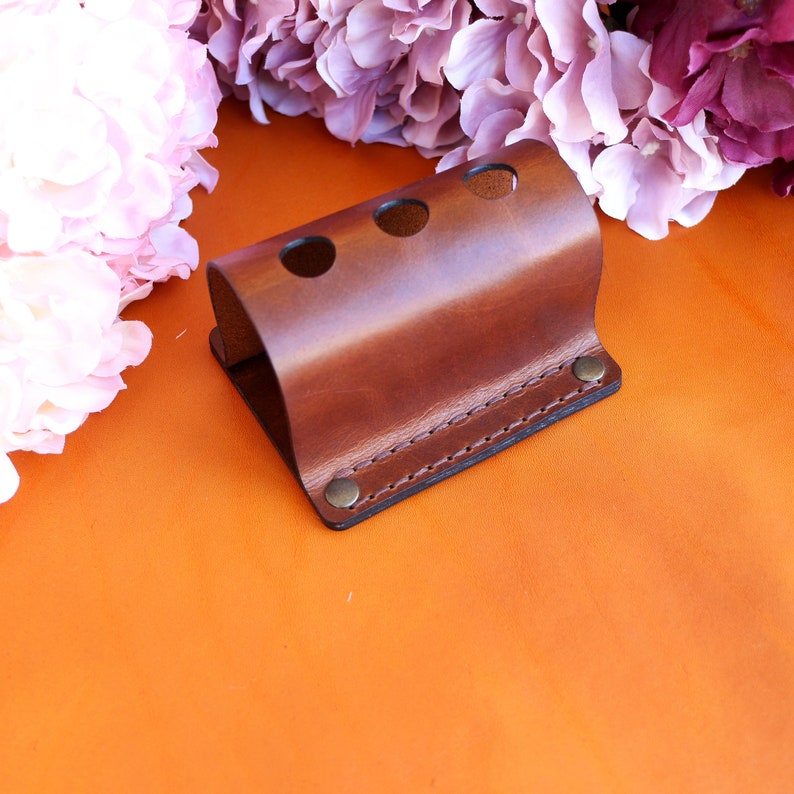 May include: A brown leather toothbrush holder with three holes. The holder is stitched with a brown thread and has a brass rivet on the bottom.