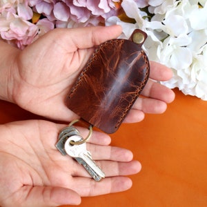 May include: A brown leather key holder with a flap closure and a brass snap. The key holder is attached to a silver key ring with three keys.