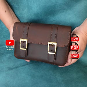 May include: A dark brown leather satchel featuring two vertical straps with gold-toned metal buckles. The bag includes text overlays reading Tutorial Video, SVG, PDF, and DXF, indicating digital file formats for crafting this item.