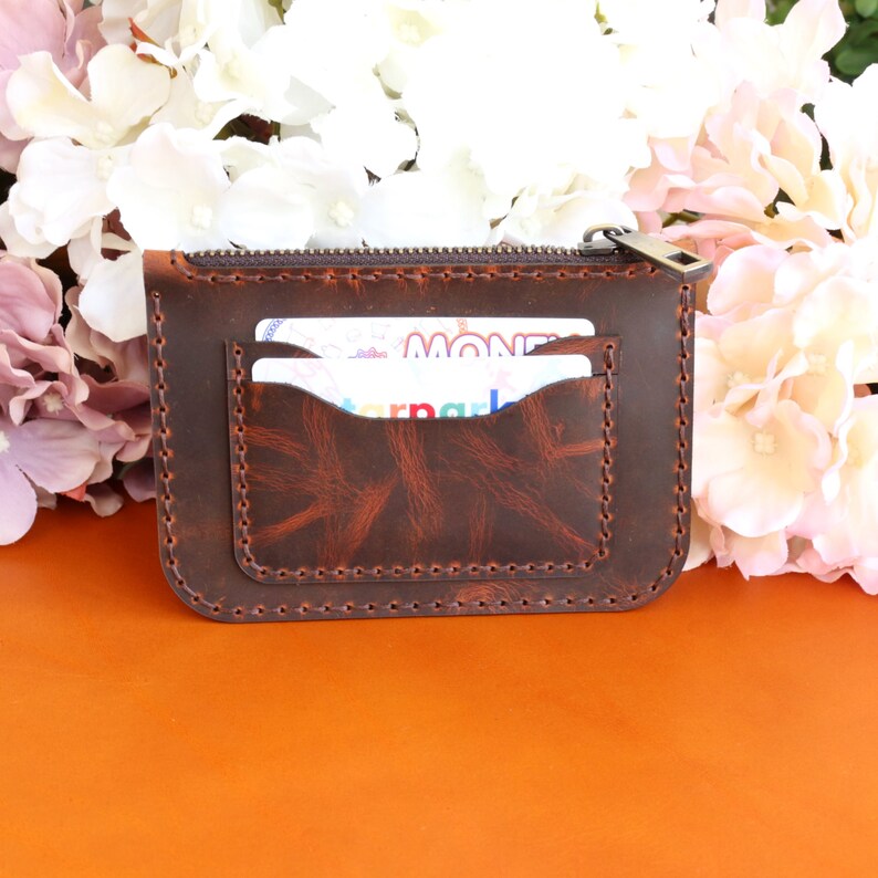 May include: A brown leather zippered wallet with a stitched design and two card slots. The wallet is lying on an orange surface.