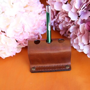 May include: A brown leather pen holder with three slots. A green pen is in one of the slots.